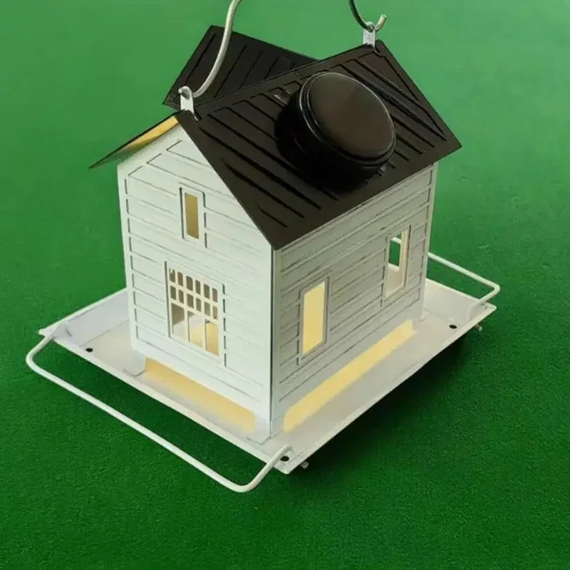 A white wooden bird feeder lantern with a dark roof and a light-colored base sits on green grass.