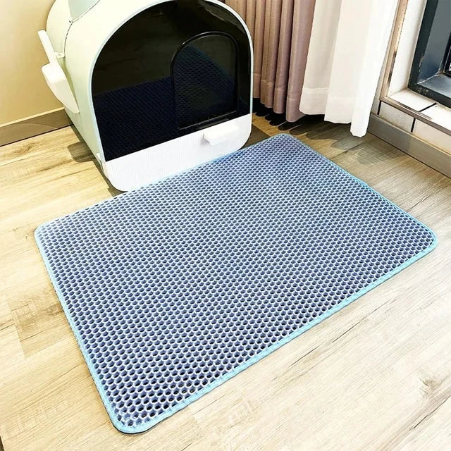 Un tapis de litière en maille bleue est placé sur un plancher en bois clair devant une boîte à litière pour animaux blanche.