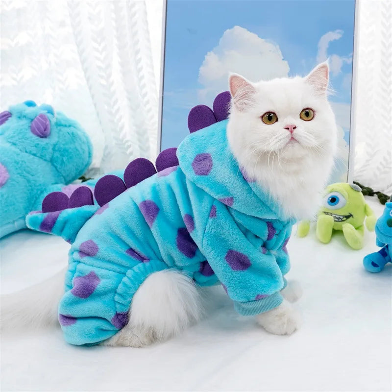 A white kitten wearing a bright blue fleece hoodie decorated with purple dinosaur shapes, sitting in front of a light blue ba