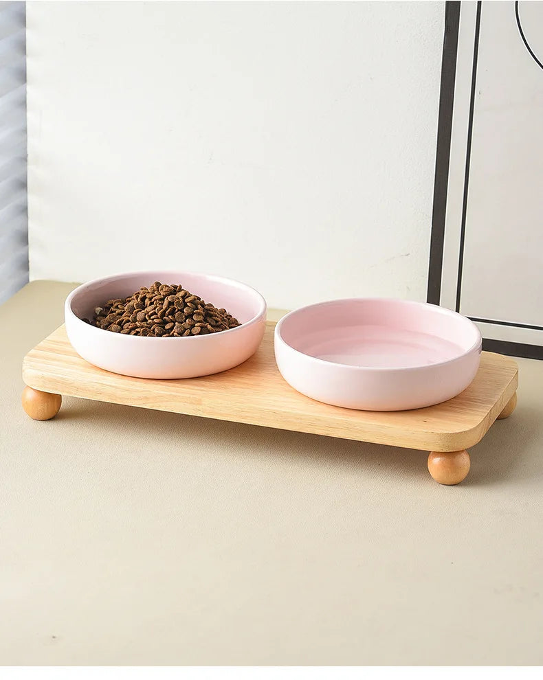 A pink ceramic cat and dog bowl is placed on a wooden stand with a pile of dry food.