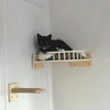 A black cat is perched on a wooden wall-mounted cat tree featuring scratching stairs and a hammock.