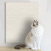 A fluffy gray and white cat is sitting in front of a textured beige mat on a wooden surface.