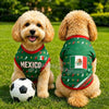 A golden doodle dog wearing a green and red soccer jersey sitting on green grass.