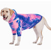 A light-colored dog wearing a tie-dye fleece hoodie with pink and blue patterns.