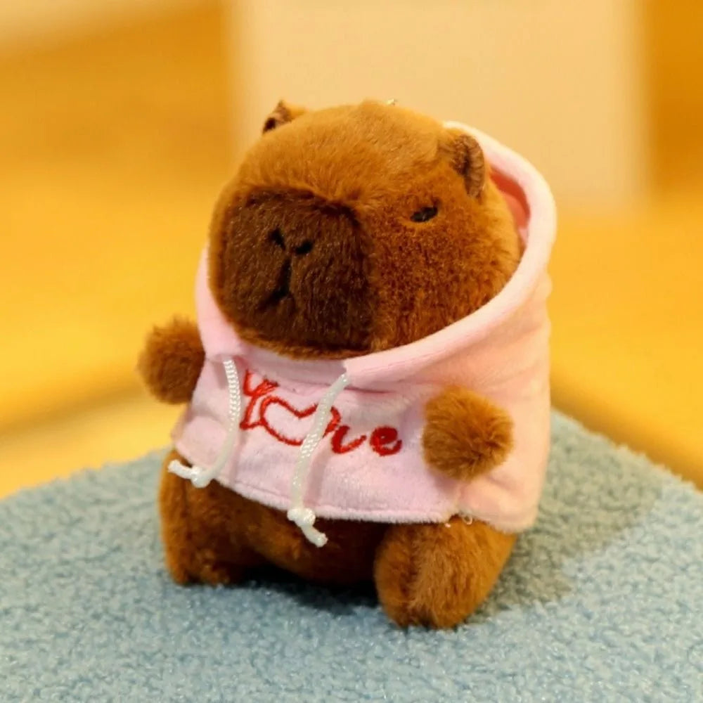 A brown capybara plush doll wearing a pink hoodie with red embroidery, sitting on a blue surface.