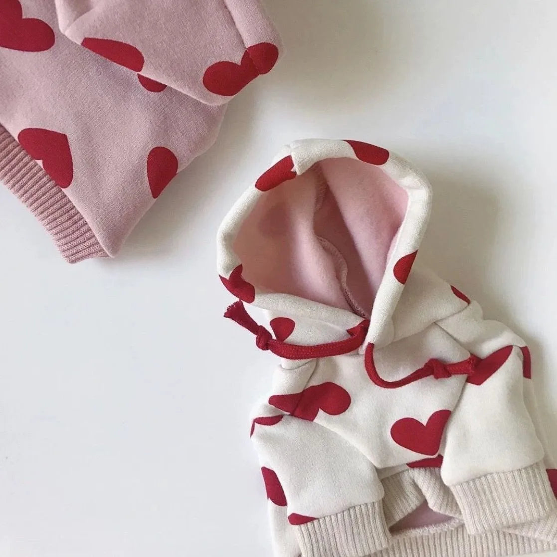 Sudadera blanca con estampados de corazón rojo y acentos rosa, doblada sobre una superficie blanca.