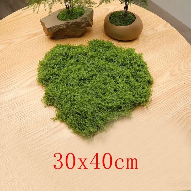 A vibrant green preserved moss clump displayed on a light wooden surface with small potted trees.
