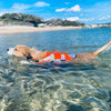 A golden dog wearing an orange reflective life jacket swimming in clear blue water near a sandy beach.